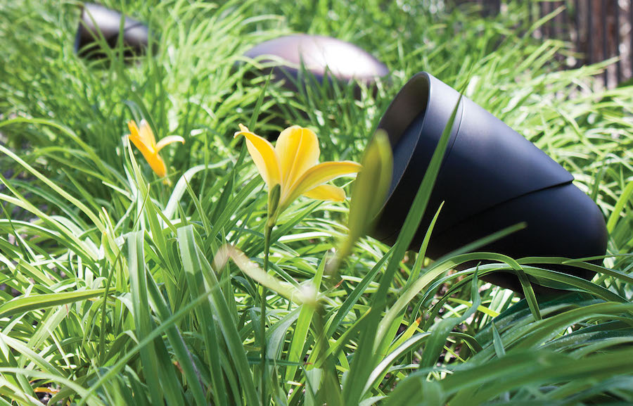 Sonance outdoor bullet speaker situated in a grassy yard with an in-ground subwoofer in the background.